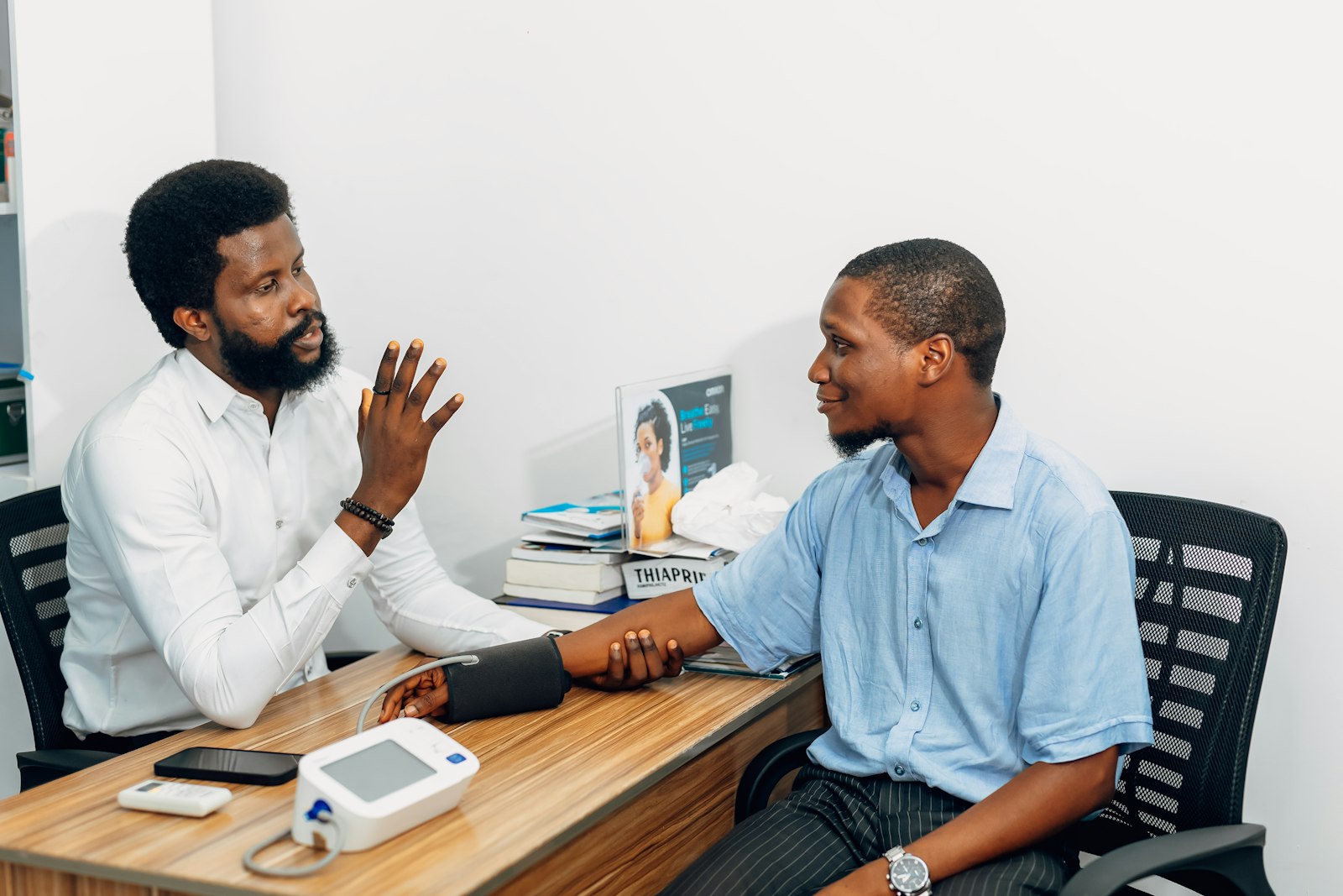 A doctor in a white shirt talking with a patient across a desk with a blood pressure monitor on it
