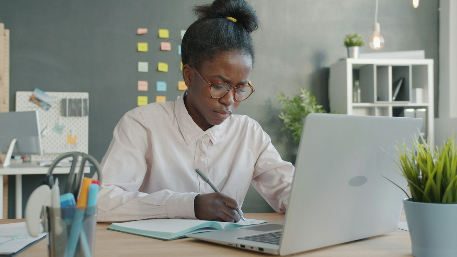 A young woman in glasses writing in a notebook at a desk beside an open laptop