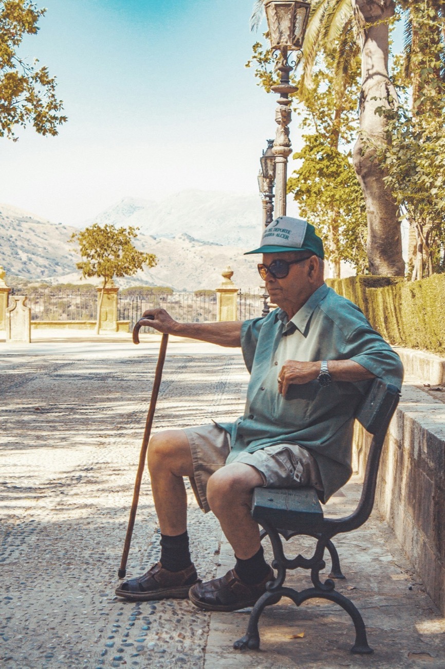 An older man in a cap and sunglasses resting on a park bench, a walking cane in his hand