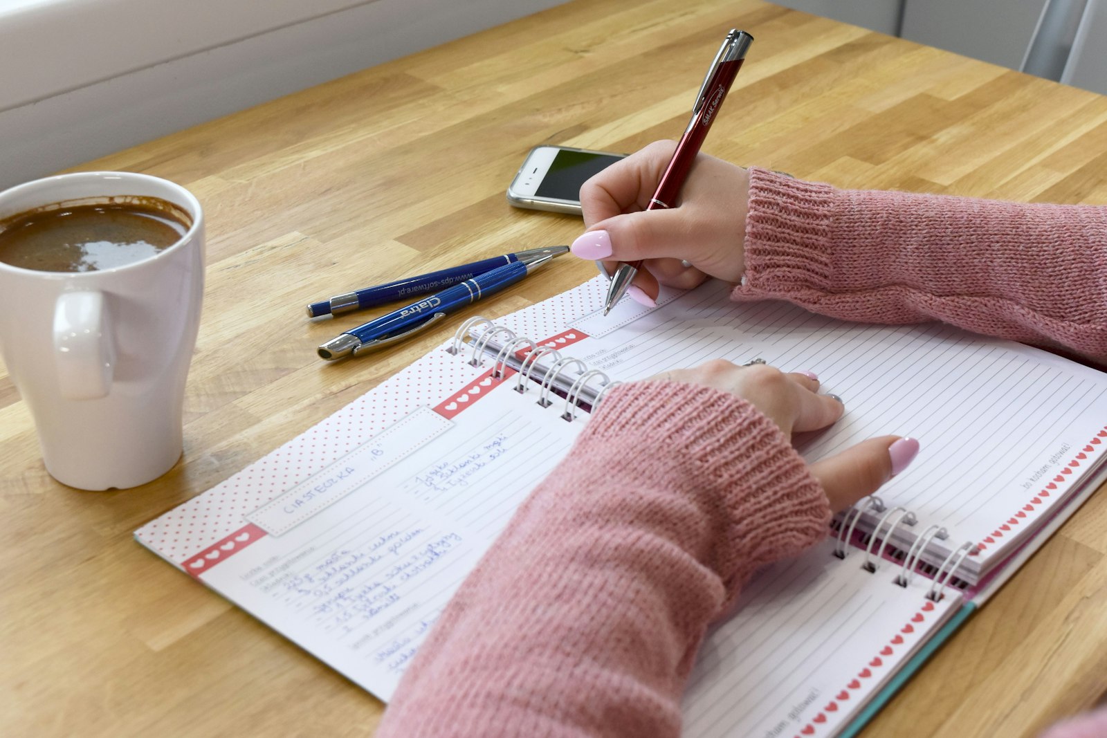 A hand writing notes in a ruled planner beside a cup of coffee