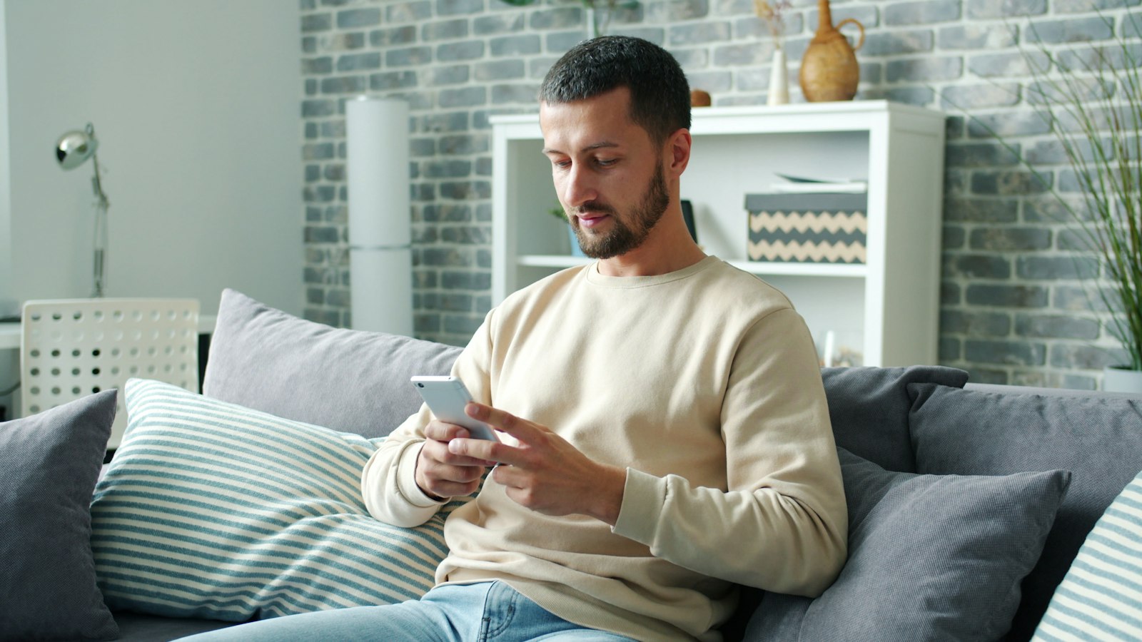 A man in a cream sweater reading a message on his phone on a sofa